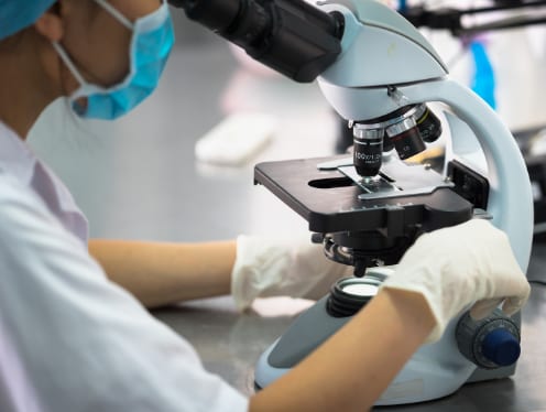 A lab technologist examining body tissue under a microscope.